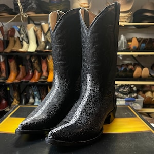 May include: A pair of black cowboy boots with a textured finish and white accents. The boots have a pointed toe and a tall shaft with decorative stitching. The boots are displayed on a black and yellow surface, with a background of other boots.
