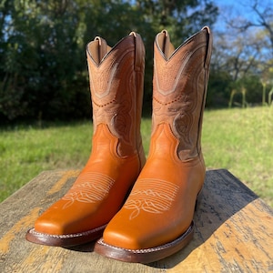May include: A pair of new, orange leather cowboy boots with a square toe. The boots have decorative stitching on the shaft and vamp, and a brown sole. The boots are displayed on a wooden surface.