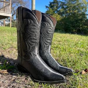 May include: A pair of black cowboy boots with detailed stitching on the upper. The boots have a textured lower section and a white stripe along the vamp. They are displayed on grass. The boots are approximately 30 cm in height.