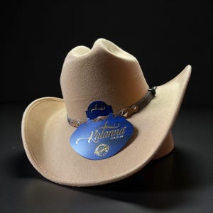 May include: A beige cowboy hat with a curved brim and a brown leather band with silver studs. A blue tag with the name "Kalanna" is attached. The hat is made of a soft material and is set against a black background.