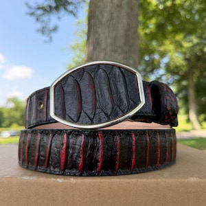 May include: A black belt with a textured, scale-like pattern and vibrant red accents between the scales. The belt features a large, oval-shaped buckle with a smooth silver-toned metal frame that encloses a matching black and red textured inlay.