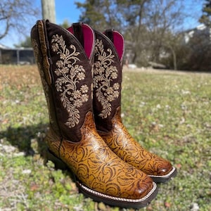 May include: A pair of brown and tan cowboy boots with floral embroidery and embossed patterns. The boots have a square toe, a dark brown sole, and a pink interior. The boots are made of leather and are suitable for country and western wear.
