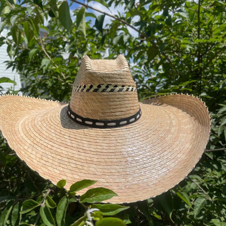 May include: A wide-brimmed straw hat with a natural beige color. The hat features a decorative band with black and white accents around the crown. The brim is large and provides ample shade. The hat is a classic accessory for sunny days.