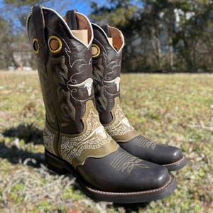 May include: A pair of brown leather cowboy boots with intricate white and olive green embroidery. The boots have a square toe, decorative stitching, and gold eyelets. The boots are set against a blurred outdoor background.