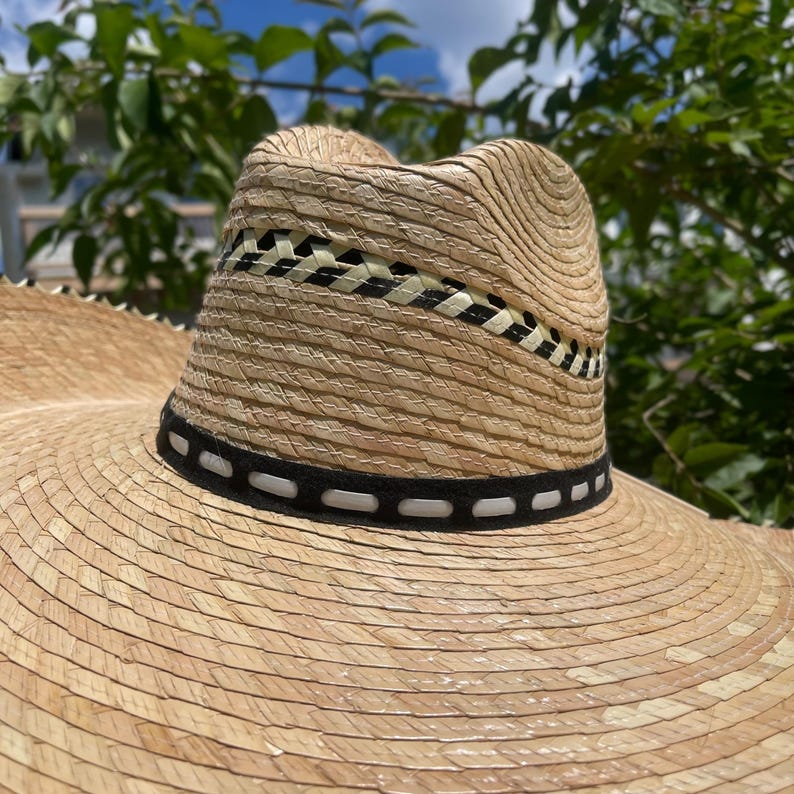May include: A wide-brimmed straw hat with a woven band featuring black, white, and beige patterns. The hat has a black band with white stitching around the crown. The hat is made of natural straw material.
