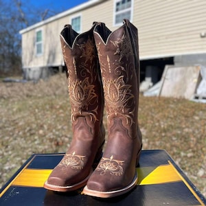May include: A pair of brown leather cowboy boots with gold floral embroidery. The boots have a square toe, a stacked heel, and a tall shaft. The boots are displayed on a black and yellow striped surface.