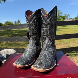 Könnte beinhalten: Ein Paar schwarze und graue Cowboy-Stiefel mit Schlangenmuster am unteren Teil und Ziernähten am oberen Schaft. Die Stiefel werden auf einer roten Box präsentiert, die ihr detailliertes Design und ihre Handwerkskunst zeigt.