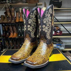 May include: A pair of brown and tan cowboy boots with floral embroidery and a square toe. The boots have a black upper with gold floral detailing and a pink interior. The lower portion of the boot is tan with a floral pattern.