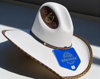 Könnte beinhalten: Weißer Cowboyhut mit braunem und goldfarbenem Rand. Der Hut hat ein braunes Lederband mit einem kleinen Juwel und einen braunen Aufnäher mit einem Hufeisen-Design. Ein blaues Etikett mit dem Text "KINGSTONE HATS" ist angebracht.