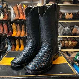 May include: A pair of black cowboy boots with a textured, scale-like pattern on the lower part and decorative stitching on the upper shaft. The boots have a square toe and a subtle blue accent. Other cowboy boots are visible in the background.