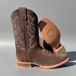 May include: Brown suede cowboy boots with decorative stitching and a square toe. The boots have a stacked heel and a leather sole. The second boot is upside down, showing the sole.