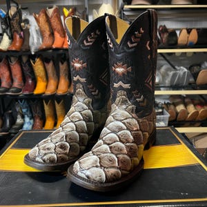 May include: A pair of cowboy boots with a distinctive design. The boots have a black upper with decorative stitching and a brown and tan patterned lower section. The boots are displayed on a black and yellow striped surface, with other boots visible in the background.