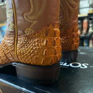 May include: Close-up of two brown cowboy boots. The boots have a textured, orange-brown alligator-like pattern on the lower part and a smooth, lighter brown leather upper. Gold stitching decorates the upper. The soles are dark brown.