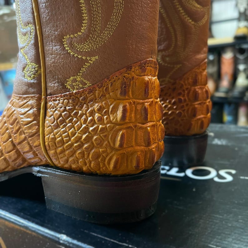 May include: Close-up of two brown cowboy boots. The boots have a textured, orange-brown alligator-like pattern on the lower part and a smooth, lighter brown leather upper. Gold stitching decorates the upper. The soles are dark brown.
