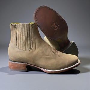 May include: A pair of brown leather cowboy boots with a square toe and stacked heel. The boots have a pull-on design with elastic side panels and decorative stitching on the vamp. The soles are brown leather with a stitched white welt.