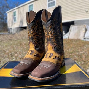 May include: A pair of brown and tan cowboy boots with detailed stitching and decorative patterns. The boots have a square toe design and are displayed on a black and yellow box. The boots are made of leather.