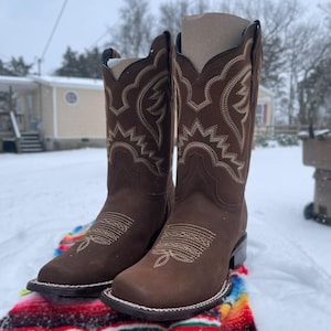 May include: A pair of brown cowboy boots with detailed light tan stitching. The boots, with a square toe, are set against a snowy background. The boots' design reflects a classic Western aesthetic.