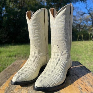 Könnte beinhalten: Ein Paar cremefarbene Cowboy-Stiefel mit einer strukturierten, krokodilartigen Musterung am Fuß und einem gestickten Design am Schaft. Die Stiefel haben eine spitze Zehenpartie und eine schwarze Sohle, vor einem natürlichen Hintergrund.