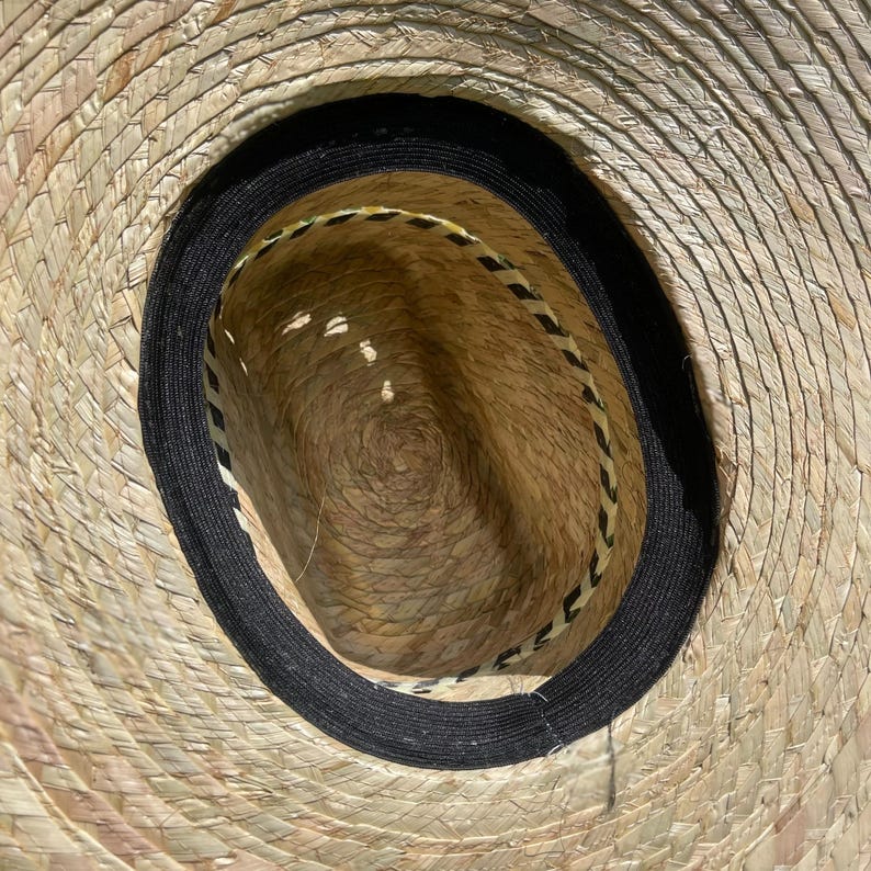 May include: A woven straw hat with a wide brim, featuring a black band around the crown and a decorative trim inside. The hat's natural tan color is contrasted by the dark band, creating a classic look.