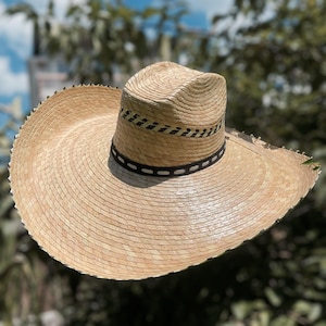 May include: A wide-brimmed straw hat with a natural beige color. The hat features a black band with white stitching around the crown and a decorative band above it. The brim has a slightly frayed edge.