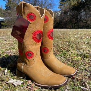 May include: Tan cowboy boots featuring red sunflower embroidery. The boots have a square toe, a stacked heel, and a tag with the text "EL GENERAL". The boots are displayed on grass. The boots are approximately 30 cm tall.
