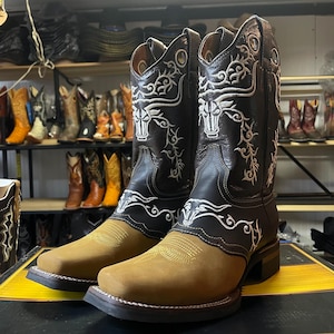 May include: A pair of brown and black cowboy boots with white decorative stitching. The boots have a square toe and a tan-coloured foot. The shaft features a bull design. Other cowboy boots are visible in the background.