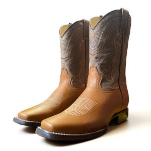 May include: A pair of tan leather cowboy boots with square toes. The boots have decorative stitching on the shaft and vamp, with a light brown and grey shaft. The soles are dark brown.