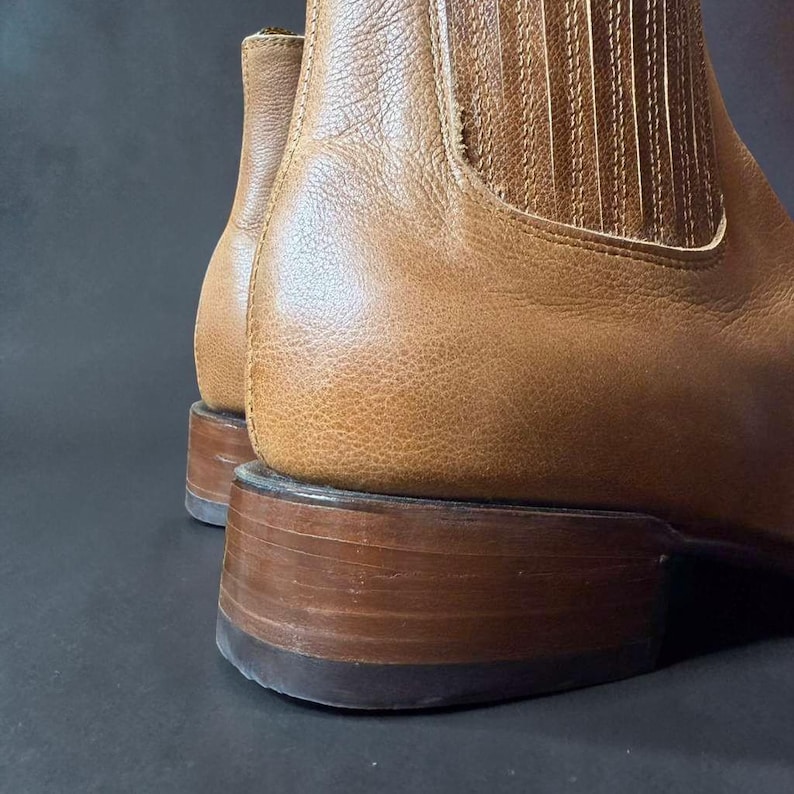 May include: Close-up of a pair of brown leather boots. The boots have a classic design with a stacked heel and elastic side panels. The leather has a textured appearance, and the boots appear to be new.