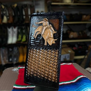 May include: A tall, rectangular leather wallet featuring a dark brown and black finish. The upper section displays a hand-tooled design of a bull rider on a bucking bull. The lower section features a woven basket-weave pattern in tan and brown.