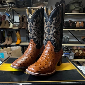 May include: A pair of brown and black cowboy boots with detailed embroidery on the black leather uppers. The boots feature a square toe and a textured leather on the lower section. Other boots are visible in the background.