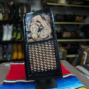 Puede incluir: Una cartera larga de cuero negro con un diseño grabado a mano de una cabeza de caballo y un sombrero de vaquero en la parte superior, con un patrón de tejido de cesta en relieve en la parte inferior. La cartera tiene forma rectangular.