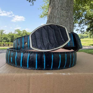 May include: A black belt featuring a textured, scale-like pattern with vibrant blue vertical stripes running along the length. The belt includes a large, oval-shaped silver-toned buckle with a matching black textured inlay in the center.