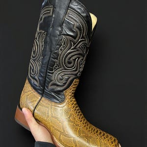 May include: A cowboy boot with a black leather upper and a tan, textured lower section. The black part has detailed white stitching. The boot has a square toe and a brown sole. The boot is held by a person.