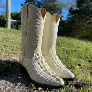 May include: A pair of off-white cowboy boots with a textured, crocodile-like pattern on the lower portion and decorative stitching on the upper shaft. The boots have a pointed toe and a black sole, set against a backdrop of green grass.