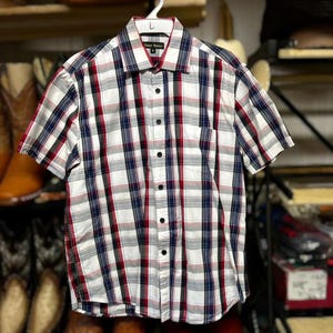 May include: A short-sleeved button-down shirt with a plaid pattern in white, red, and navy blue. The shirt has a chest pocket and is hanging on a white hanger. The shirt is made of a lightweight fabric.