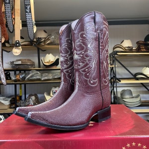 May include: Burgundy cowboy boots with silver embroidery. The boots have a pointed toe and a square heel. The background shows a shop with belts and hats on display.