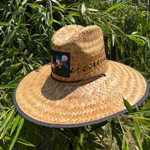 Op de afbeelding: Een strooien hoed met brede rand en een natuurlijke beige geweven textuur, voorzien van een zwarte stoffen bies langs de rand. Een vierkante patch op de voorkant toont een kleurrijke haan-afbeelding. Een dun zwart koord omcirkelt de kroon.