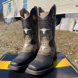May include: A pair of brown leather cowboy boots with decorative stitching and a bull head design. The boots have a square toe and a stacked heel. The shaft features a decorative design in white and olive green.