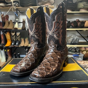 May include: A pair of brown leather cowboy boots with a textured, scale-like pattern on the foot and a decorative stitched design on the shaft. The boots have a pointed toe and a stacked heel, displayed on a box.