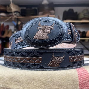 May include: Black leather belt with a large oval buckle featuring a brown embroidered bull. The belt has brown and black geometric embroidery and a matching bull design. White stitching outlines the buckle and belt.