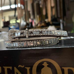 Könnte beinhalten: Zwei gestapelte braune Ledergürtel mit silbernen rechteckigen Strasssteinen und einer silbernen Schnalle. Die Gürtel sind mit Reihen funkelnder Strasssteine verziert, was ein glamouröses Accessoire ergibt. Die Schnalle ist ebenfalls mit Strasssteinen besetzt.