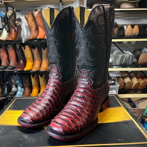 May include: A pair of cowboy boots with a red and black snakeskin pattern. The boots have a black leather upper with decorative stitching and a red sole. Other boots are visible in the background.