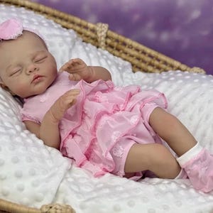 Może przedstawiać: Realistyczna lalka w różowej sukience z falbanami i pasującą opaską z kokardą. Lalka leży na białym, teksturowanym kocu w wiklinowym koszu. Lalka ma zamknięte oczy i nosi różowe skarpetki.