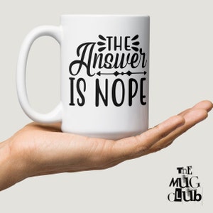 May include: White ceramic coffee mug with a black design that says "The Answer Is Nope" with an arrow pointing to the right.
