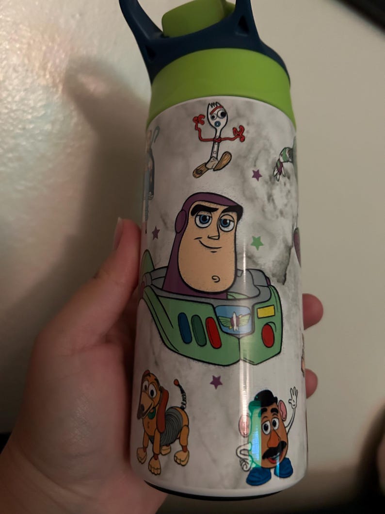 May include: A stainless steel water bottle featuring characters from the animated film Toy Story. The bottle has a white marble-like background with images of Buzz Lightyear, Forky, Slinky Dog, and Mr. Potato Head. The lid is green and blue.