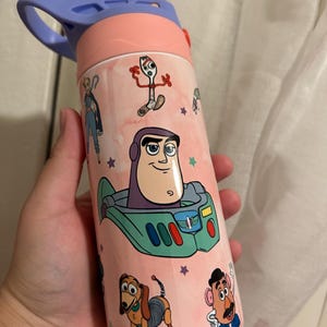 May include: A pink and blue water bottle decorated with characters from Toy Story. The bottle features Buzz Lightyear, Forky, Slinky Dog, and Mr. Potato Head. The lid is light blue and has a handle. The bottle is designed for children.
