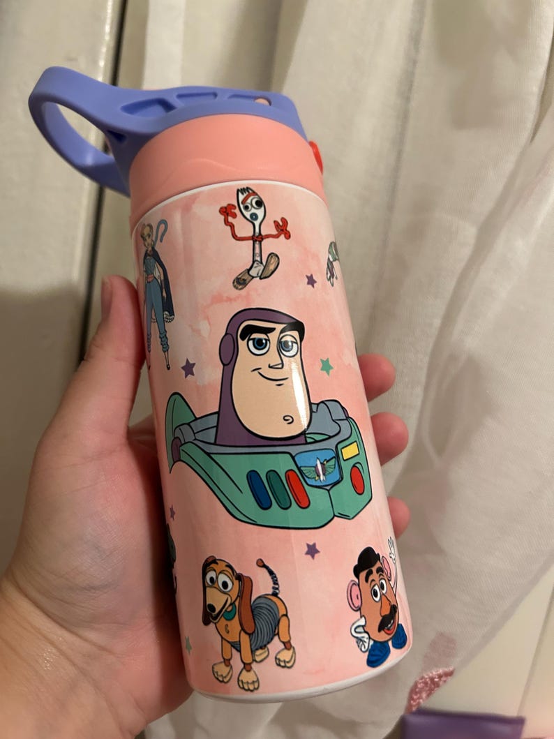 May include: A pink and blue water bottle decorated with characters from Toy Story. The bottle features Buzz Lightyear, Forky, Slinky Dog, and Mr. Potato Head. The lid is light blue and has a handle. The bottle is designed for children.