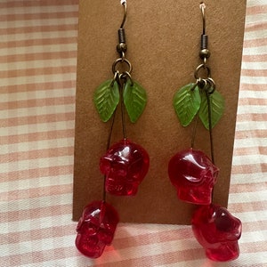 May include: A pair of dangle earrings featuring red skull charms. The earrings have green leaf accents and are suspended from bronze-colored hooks. The skulls are a translucent red color.
