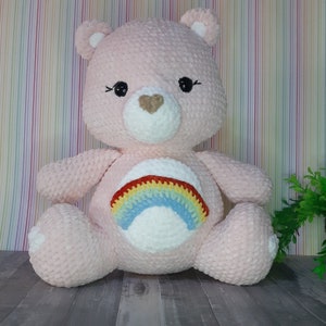 Puede incluir: Un oso de peluche rosa tejido a crochet con un arcoíris bordado en el pecho. El oso tiene una nariz blanca en forma de corazón y ojos negros.