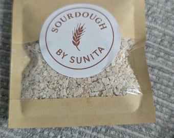 May include: A small, rectangular, light brown paper pouch with a clear plastic window containing dried, beige, flaky sourdough starter. A circular white sticker on the front features a wheat stalk icon and the text SOURDOUGH BY SUNITA.